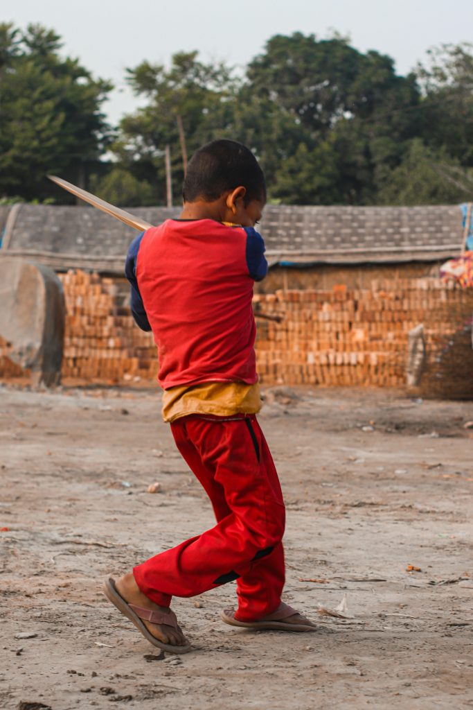 A young boy enjoys playing cricket on a rustic street, wearing a vibrant red shirt.